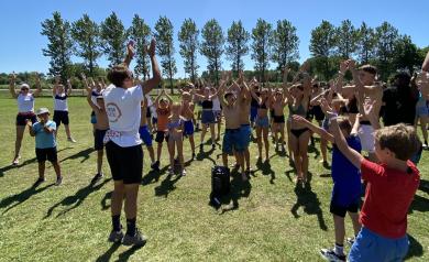 Animateur du mini club guidant un groupe d’enfants lors d’une sortie sportive aux terrains multisports, tous vêtus de tenues décontractées, en déplacement dynamique sous un ciel clair, entourés de buts, paniers et équipements sportifs en plein air.