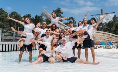 Photo de groupe des animateurs du camping Le Vieux Port saison 2025, posant souriants et en tenue décontractée au parc aquatique, entourés de structures aquatiques colorées, toboggans et bassins, sous un ciel clair d’été.