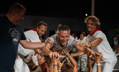 Homme levé sur les bras d’autres campeurs pendant la soirée Féria au Parvis de l'Arena, la plus grande et merveilleuse salle événementielle des Landes, sous un ciel nocturne animé, dans une ambiance festive et conviviale.