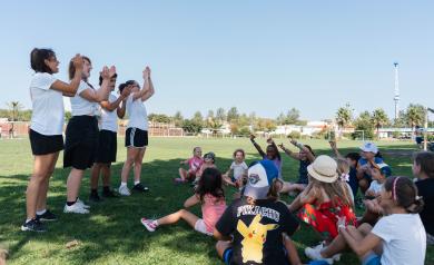 Animateurs du mini club donnant des consignes aux enfants rassemblés sur un terrain de football ombragé, dans une atmosphère calme et attentive, avant le début de la prochaine activité sportive, avec arbres fournissant de l’ombre et équipement sportif visible en arrière-plan.