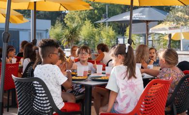 Enfants du mini club réunis sous des parasols colorés, savourant leur repas avec joie et bonne humeur, partageant rires et sourires dans une ambiance conviviale et détendue au cœur du camping, entourés de tables et chaises adaptées à leur taille.