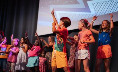 Spectacle hebdomadaire des enfants au camping Le Vieux Port, tous les vendredis soir dans la salle l'ARENA, mettant en vedette les jeunes stars des familles qui montent sur scène pour danser et jouer devant un public joyeux de parents et vacanciers, dans une atmosphère festive et chaleureuse.