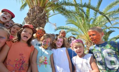 Enfants du mini club du camping Le Vieux Port, fiers et souriants après une activité de maquillage de masques, posant devant une vue sur de grands palmiers et un ciel bleu lumineux, dans une atmosphère joyeuse et accueillante au cœur du club enfants.