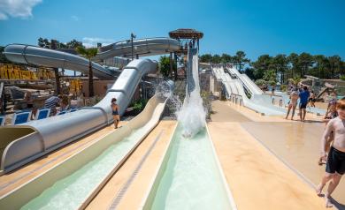Sorties de plusieurs toboggans aquatiques au parc aquatique XXL du camping Le Vieux Port dans les Landes, avec des couloirs d'eau clairs dévalés par les vacanciers, entourés de structures colorées comme le Kamikaze, le Black Hole et le Pentagliss, dans un espace vaste et thématique inspiré de la piraterie, sous un ciel bleu et une végétation luxuriante typique de la pinède landaise.