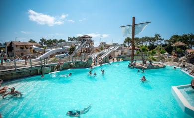 Grande piscine en forme de bateau située sur la plage du parc aquatique XXL du camping Le Vieux Port dans les Landes, avec un jacuzzi chauffé adjacent, entourée de sable fin et palmiers, offrant un cadre idyllique pour la détente au bord de l’océan Atlantique, sous un ciel bleu lumineux et une ambiance paradisiaque.