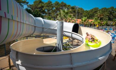 Un papa et son fils souriants partageant un moment de joie sur une bouée double en descente dans le toboggan à virages du parc aquatique XXL du camping Le Vieux Port dans les Landes, entourés d’eaux claires, glissant à toute vitesse à travers les courbes du toboggan dans une ambiance familiale et ludique.
