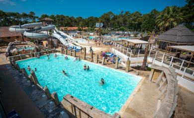Vue panoramique de la grande piscine extérieure du parc aquatique XXL du camping Le Vieux Port dans les Landes, avec un bassin spacieux et tempéré accueillant des cours de natation, des compétitions de water-polo, et diverses activités aquatiques, entouré de palmiers et de toboggans colorés, dans un environnement familial et convivial au cœur d’une pinède en bord de mer.