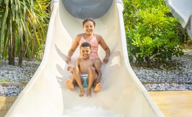 Deux jeunes souriants en maillot de bain sortant ensemble du toboggan aquatique à grande vitesse du parc aquatique XXL du camping Le Vieux Port, entourés d'eau claire et de jets d'eau, capturant un moment de joie et d’amusement durant une chaude journée d’été dans un cadre naturel de pinède en bord de mer.