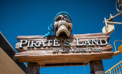 ntrée colorée et thématique de Pirate's Land, l’aire d’aquasplash du camping 5 étoiles Le Vieux Port dans les Landes, avec des jeux d’eau ludiques et sécurisés adaptés aux enfants, incluant toboggans petits jets d’eau, et une grande structure en forme de crâne de pirate déversant de l’eau, entourée de palmiers dans un décor de piraterie joyeux et vibrant.