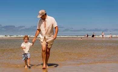 Un père et son fils debout sur le littoral landais, les pieds dans l'eau claire et calme à marée basse, entourés d’une étendue de sable fin et de petites baïnes, sous un ciel dégagé, avec l’horizon maritime à perte de vue, dans un cadre naturel typique de la côte landaise marqué par la lumière douce, la sérénité et la beauté du bord de mer.