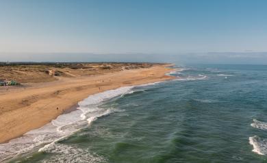 Vue aérienne prise par drone en inclinaison à 45° vers le bas, dévoilant l’océan Atlantique à droite aux eaux bleu turquoise avec de douces vagues, et la plage de sable fin à gauche bordée de dunes et de végétation typique de la pinède landaise, sous un ciel clair, offrant un panorama spectaculaire et naturel du littoral.