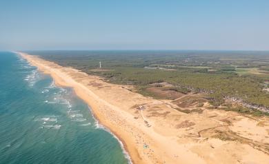 Vue aérienne prise par drone avec une inclinaison de 45° vers le bas, montrant l’océan Atlantique à gauche avec ses eaux bleu turquoise et ses vagues douces, et la plage de sable fin à droite bordée de dunes et de végétation typique de la pinède landaise, sous un ciel clair et lumineux, offrant un panorama naturel et spectaculaire du littoral landais.