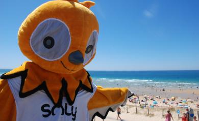 Soly, la mascotte joyeuse du camping Le Vieux Port dans les Landes, posant avec les vacanciers souriants sur la plage de sable fin aux eaux turquoise de l’océan Atlantique, sous un ciel lumineux, incarnant la convivialité et le bonheur d’un séjour inoubliable en bord de mer dans un cadre naturel préservé et familial.