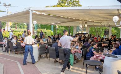 Serveurs en tenue professionnelle servant des boissons fraîches et des apéritifs à des vacanciers souriants assis aux tables en terrasse du bar Le VP, au camping Le Vieux Port dans les Landes. L’ambiance est conviviale et détendue, avec des clients profitant du soleil sur une grande terrasse en bois, entourée de pins, dans un cadre chaleureux et festif en bord de mer.