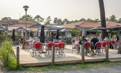 Nouvelle terrasse spacieuse et moderne du bar Le VP au camping Le Vieux Port dans les Landes, avec des tables en bois clair et chaises confortables disposées sous une pergola offrant de l’ombre, entourée de plantes et de lumières d’ambiance, créant un espace convivial et élégant où les vacanciers peuvent se détendre, savourer un verre ou un repas en profitant de la douceur du climat landais.