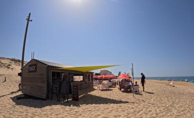Snack bar situé directement sur la plage, les pieds dans le sable, au camping Le Vieux Port dans les Landes, accueillant des vacanciers assis à des tables, profitant de boissons fraîches et de snacks savoureux dans une atmosphère conviviale et détendue au bord de l’océan Atlantique. L’espace est entouré de la dune, avec vue sur la plage et accès direct par un chemin sécurisé depuis le camping.