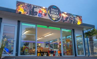 Vue extérieure de la salle de jeux vidéo et d’arcade du camping Le Vieux Port dans les Landes, avec un espace spacieux et convivial, équipé de nombreuses bornes d’arcade colorées, de machines de jeux rétro, de simulateurs de voitures, de billards, d’un flipper, de baby-foot, et de consoles modernes. La salle est animée par des vacanciers de tous âges, certains en train de jouer, d’autres en groupe ou en famille, dans une ambiance familiale et dynamique, avec un décor moderne et lumineux, idéale pour s’amuse