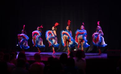 Spectacle cabaret French Cancan présenté à l’Arena du camping Le Vieux Port, avec des danseuses en costumes traditionnels colorés et à jupes volantes, exécutant des figures emblématiques sur scène devant un public enthousiaste. La scène est illuminée par des jeux de lumière dynamique qui mettent en valeur l’énergie et la vivacité de la danse, dans un cadre élégant et festif de la plus grande salle de spectacles des Landes, offrant une expérience culturelle et divertissante typique des cabarets français.
