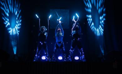 Spectacle dynamique à l’Arena du camping Le Vieux Port avec trois danseurs évoluant sur une scène éclairée, portant des costumes futuristes et manipulant des accessoires LED colorés qui créent des jeux de lumière hypnotiques. La scène spacieuse et équipée de matériel professionnel met en valeur la chorégraphie synchronisée et l’énergie des artistes, dans une ambiance moderne et immersive, captivant un public nombreux dans la plus grande salle de spectacles des Landes, offrant une belle expérience visuelle.
