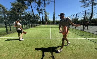 Des joueurs disputant un match de padel sur un terrain clôturé de verre et grillage, sous un ciel dégagé. Quatre participants en tenues de sport, équipés de raquettes spécifiques au padel, sont en pleine action, se concentrant sur la balle qui vole entre eux. Le terrain, doté d'un sol en gazon artificiel, est entouré d'un public limité ou d'un cadre naturel, avec une ambiance sportive et conviviale, où l'agilité, la rapidité et la stratégie sont essentielles au jeu.