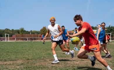 Des joueurs en pleine action sur le terrain multisports du camping, disputant un match de rugby sur un gazon bien entretenu. Plusieurs sportifs en tenues adaptées au rugby sont visibles, certains courant avec le ballon ovale, d’autres tentant de plaquer ou de se positionner pour recevoir la passe. L’ambiance est dynamique et sportive, avec un ciel clair en arrière-plan et une équipe concentrée sur le jeu collectif dans un cadre naturel et verdoyant typique d’un camping offrant des installations multisports.
