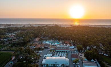 Vue aérienne panoramique du camping Le Vieux Port au crépuscule, montrant un vaste domaine avec des emplacements de mobil-homes et tentes entourés de pins majestueux et de chemins sinueux. Au loin, l’horizon clair dévoile un coucher de soleil flamboyant aux teintes orangées et rosées, baignant le paysage d’une lumière douce et chaleureuse. Le ciel se pare de nuages légers, tandis que la mer ou la forêt environnante se dessine à l’horizon, créant une atmosphère paisible et magique typique d’une soirée d’été.