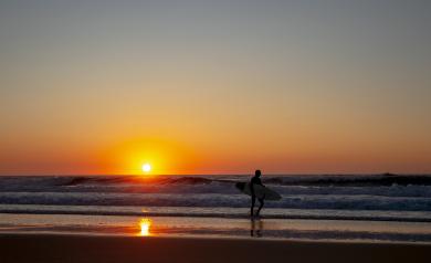 Un surfeur marche calmement dans l'eau peu profonde avec sa planche de surf sous le bras, les pieds effleurant la surface de la mer au coucher du soleil. Le ciel est peint de teintes chaudes d’orange, de rose et de violet, illuminant doucement la silhouette du surfeur. L'eau reflète les couleurs du ciel, créant une atmosphère paisible et contemplative, tandis que le soleil descend lentement vers l’horizon. La scène capture la beauté naturelle, la sérénité et la connexion intime entre le surfeur et l’océan.