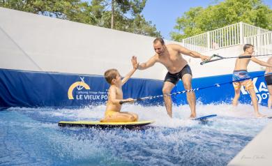 Un père et son jeune fils se tapant la main avec enthousiasme, debout dans l’eau peu profonde du bassin de surf à la Vieux Port Surfing Wave, dans le parc aquatique. Le père porte un maillot de bain coloré et un sourire large, tandis que le fils, également en maillot de bain, montre une expression de joie et de concentration. En arrière-plan, on voit la grande machine à vagues et d’autres activités aquatiques, sous un ciel bleu clair et un soleil brillant. La scène respire la complicité, le plaisir.