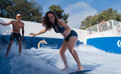 Une femme participante à l’animation surfing contest adultes sur la Vieux Port Surfing Wave du parc aquatique du camping Le Vieux Port. Elle est debout en équilibre sur sa planche de surf artificielle, concentrée et souriante, vêtue d’une combinaison de sport adaptée. Autour d’elle, l’eau en mouvement simule des vagues dynamiques créées par la machine à surf WaveMotion, dans un environnement sécurisé et moderne. En arrière-plan, on distingue d’autres participants et animateurs encadrant l’activité.
