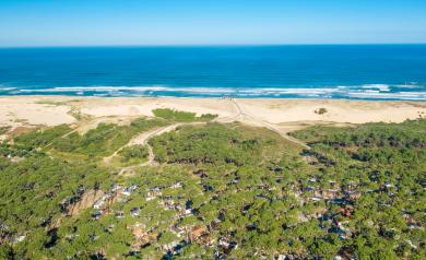 Vue panoramique aérienne prise par drone du camping, montrant les emplacements verts et les structures du camping en premier plan, avec un vaste horizon bleu de l’océan Atlantique s’étendant à l’arrière-plan sous un ciel clair et lumineux