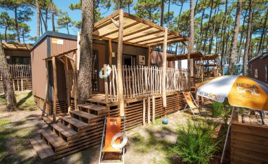 Vue extérieure d'un locatif moderne et élégant avec façade en bois chaleureux, grandes baies vitrées ouvertes sur un jardin verdoyant, terrasse en bois avec mobilier design pour profiter des beaux jours, ciel bleu lumineux en arrière-plan, ambiance accueillante et confortable invitant à la découverte et à la détente.