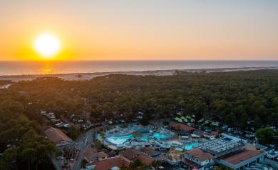 Photo aérienne prise par drone au coucher du soleil, avec vue panoramique sur l'horizon où le soleil se couche sur l'océan Atlantique, lumière dorée reflétée sur les vagues, camping visible en bas de l'image, entouré de verdure, avec son grand parc aquatique remarquable comprenant piscines et toboggans, atmosphère paisible et chaleureuse typique des heures dorées.