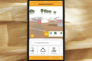 le téléphone est sur un tissu doré avec la vision 3D des bâtiments et arbres du camping pour présenter tous les services rendus par ce guide numérique avec fonction recherche