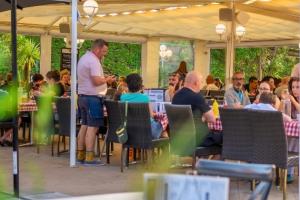 Vue sur la brasserie du VP depuis les buissons avec au premier plan une table vide et la carte du menu en dépliant ainsi que sur la majeure partie de la photo des clients et serveurs en appétit