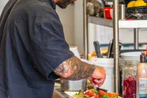 Assiette en préparation à la cuisine pro par un homme avec casquette, salade fraîche et boissons fraîches posées près du bar, ambiance estivale et familiale
