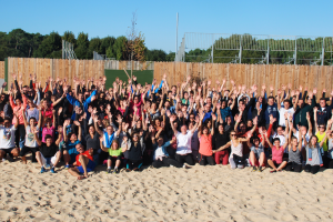 Groupe d'environ cent personnes réunies en extérieur lors d'une activité de team-building, levant toutes les mains en l'air en signe de participation et d'enthousiasme, avec des sourires, des tenues décontractées