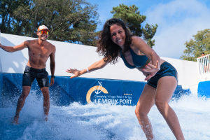 Jeune femme souriante portant un maillot de bain, en position debout sur une planche de surf à l’intérieur d'une zone d'entraînement, utilisant une machine à vagues pour pratiquer le surf