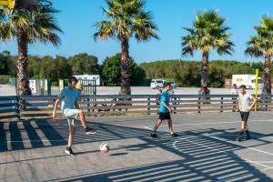 Trois enfants en pleine partie de football sur le city stade du camping, courant et tirant dans le ballon sur un terrain multisports extérieur, dans une ambiance joyeuse et dynamique.