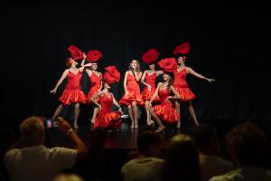 Scène de cabaret animée avec sept artistes féminines vêtues de costumes rouges éclatants, comprenant une chanteuse élégante et six danseuses en talons hauts. Les danseuses manient des froufrous rouges et des roses dans une chorégraphie enjouée, tandis que le public admiratif est visible au premier plan sous la lumière chaleureuse du spectacle. Ambiance festive et glamour typique du cabaret.