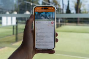 Main gauche tenant un téléphone affichant un système de guidage, prise au bord d’un court de tennis padel dans le camping, avec le planning RDV pour se rencontrer en arrière-plan.