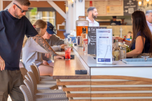 Îlot central du bar de la brasserie du VP situé dans un camping convivial des Landes, composé d’un comptoir moderne en bois clair, de chaises hautes autour de l’îlot, d’une surface élégamment décorée avec présentoirs de verres, machines à café professionnelles et tireuses à bière, avec en arrière-plan des étagères garnies de bouteilles et une lumière tamisée créant une ambiance chaleureuse et accueillante, idéale pour se retrouver entre amis ou en famille.