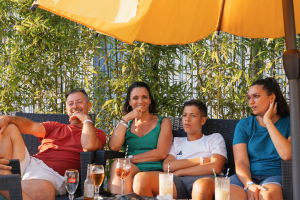 Plusieurs personnes installées confortablement sur des canapés de jardin design et bas, disposés autour de petites tables sur la terrasse parvis du bar de l’Arena, profitant d’une ambiance conviviale et détendue en plein air, entourés de coussins moelleux et de mobilier lounge, discutant ou partageant un verre dans un environnement moderne et accueillant, avec la lumière naturelle qui met en valeur l’élégance et la qualité du mobilier d’extérieur.