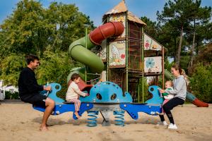 Photo d'une aire de jeux pour enfants de 5 à 12 ans dans un parc de camping, avec un sol en sable. On voit un jeu d’équilibre à ressorts occupé par un père et son garçon d’un côté, et une maman avec sa petite fille en bas de l'autre côté, tous deux en train de jouer et de s'amuser ensemble
