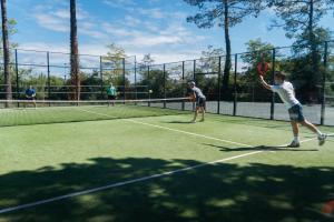 Quatre personnes jouent un match de padel sur le terrain du camping Le Vieux Port, avec deux jeunes et deux hommes d'âge mûr, tous souriants et concentrés, entourés de la structure en verre et en métal du terrain, dans une ambiance conviviale et sportive