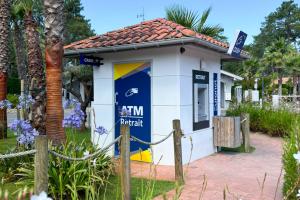 Distributeur automatique de billets situé dans un camping, entouré de palmiers et d’une végétation luxuriante, illuminé par un soleil éclatant de journée claire. Le dispositif est installé sur une zone pavée propre, reflétant un environnement naturel et agréable typique d’un lieu de vacances en plein air.