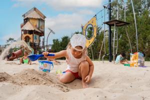 Enfant d'environ 7 ans souriant et jouant dans l'immense bac à sable de l'aire de jeux Kids du camping Le Vieux Port à Messanges, réservé aux 6-12 ans. Le garçon porte un t-shirt coloré et un short, ses pieds nus s’enfoncent dans le sable doux tandis qu’il grimpe sur le bateau bleu emblématique, entouré de balançoires, toboggans et portiques adaptés à son âge. Au fond, d'autres enfants s’amusent et découvrent les installations ludiques sous la surveillance de leurs parents, dans un cadre naturel
