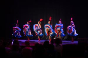 Spectacle cabaret French Cancan présenté à l’Arena du camping Le Vieux Port, avec des danseuses en costumes traditionnels colorés et à jupes volantes, exécutant des figures emblématiques sur scène devant un public enthousiaste. La scène est illuminée par des jeux de lumière dynamique qui mettent en valeur l’énergie et la vivacité de la danse, dans un cadre élégant et festif de la plus grande salle de spectacles des Landes, offrant une expérience culturelle et divertissante typique des cabarets français.