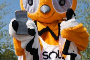 Mascotte Soly tenant un smartphone affichant l’application RESASOL, mettant en avant la réservation digitale intégrée des activités partenaires du camping Le Vieux Port, facilitant l’organisation et la gestion des loisirs