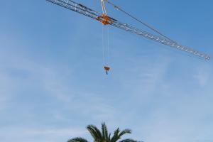 Vue depuis le sol d’une grue de chantier imposante au-dessus d’un palmier, avec le ciel bleu en arrière-plan, illustrant un chantier en cours avec matériel de construction et végétation