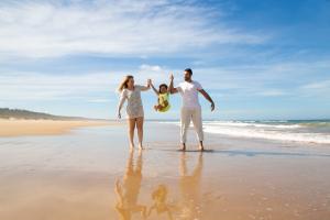 Un couple de parents à la plage tenant leur enfant par les bras pour le soulever du sol, tous trois en pleine séance de fous rires et de joie familiale, sur un sable doré avec la mer en arrière-plan sous un ciel clair.